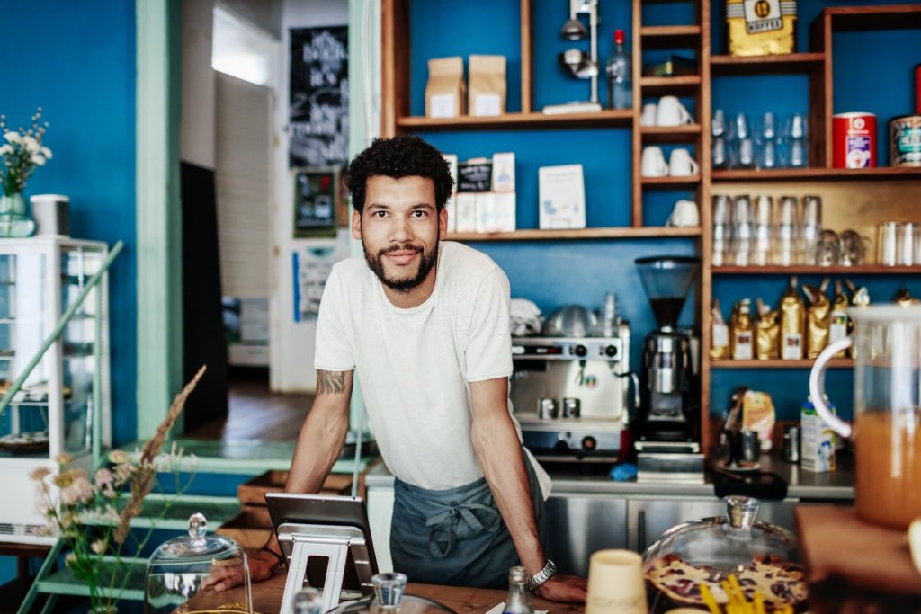 A young barista is smiling as he leans on the counter in his modern, colourful coffee shop.